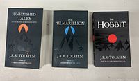 Three Tolkien books arranged side by side showing front covers: Unfinished Tales, The Silmarillion, The Hobbit.