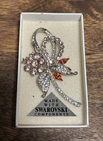 Close-up of the Swarovski crystal brooch inside a white box showing the clear, pink, and orange crystals in a floral ribbon design.