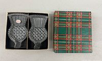 Pair of clear glass trinket dishes shaped like thistles, placed inside an open tartan-patterned box, showing both dishes side by side.