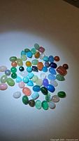 Photo showing approx. 50 polished cabochon gems of various colors and types including blue, green, pink, orange, and white stones scattered on a neutral background under direct light.