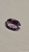 Photo of natural oval cut amethyst gemstone on a light grey surface, showing the faceted cut and color variations from pink to purple.