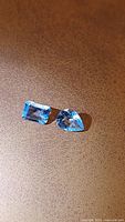 Two light blue natural aquamarine gemstones on a brown textured surface; one rectangular cut and one pear-shaped cut.