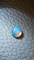 Close-up photo of a round cabochon natural Ethiopian black opal showing multicolored play of color on a black textured background.