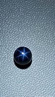 Close-up photo of a round blue star sapphire cabochon showing a six-ray star pattern on dark blue surface under light.