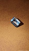 Close-up image of a natural Brazilian aquamarine gemstone, emerald cut, light blue color, placed on a brown textured surface.
