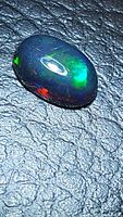 Close-up photo showing oval cabochon cut 1.38ct Ethiopian black opal with multicolored flashes on textured dark surface.