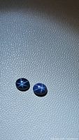 Two natural oval cabochon blue star sapphires showing clear six-rayed star reflections on a textured surface, photographed under bright light to highlight the asterism effect.
