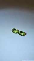Pair of apple green natural peridot gemstones, oval cut, shown on a light background to highlight color and clarity.