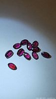 Close-up image of 11 deep purple-pink oval cut Rhodolite garnet gemstones scattered on flat surface under direct light, showing clarity and faceting.