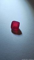Natural red cushion cut ruby gemstone placed on a white surface with light casting shadow, showing rich red color and stone transparency.