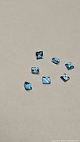 Seven natural Swiss blue topaz gemstones on a neutral background, showing multiple cuts and deep blue color.