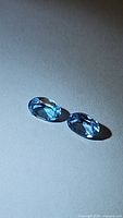 Pair of natural Swiss blue topaz gemstones, oval cut, shown against neutral surface under directional lighting highlighting facets and color.