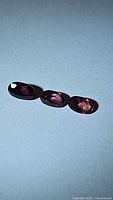 Photo showing three dark purple oval cut sapphires arranged in a row on a light blue background, displaying their rich color and clarity.