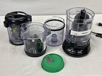 Photo showing four clear plastic food processing containers and one green lid with blades, arranged around the Ninja base unit.