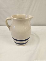 Full view of ceramic pitcher showing off-white body with 2 blue horizontal stripes and handle with spout