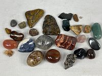 Collection of 21 assorted polished stones and minerals on white cloth background, showing variety in shape, color, and size.