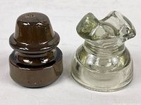 Photo showing both vintage electrical insulators, porcelain one chocolate brown and glass one clear with markings, side by side for scale and condition.
