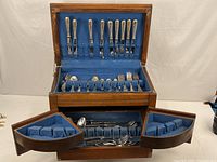 Full view of wooden chest open, showing blue felt-lined compartments with vintage silverplate and stainless flatware inside.