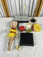Photo showing Betty Boop doll in yellow dress with red accents, various gift boxes, yellow hat, small Betty Boop figurine, and display stands arranged on a table.