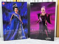 Two boxed Bob Mackie Jewel Essence Collection Barbie dolls, Sapphire Splendor and Amethyst Aura, showing box fronts with doll images and signatures.