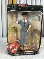 Full view of the Barbie doll in packaging showing the I Love Lucy Collector Edition box, minor damage visible on box edges, doll wearing black and white houndstooth dress and posed with arm raised holding a spoon.