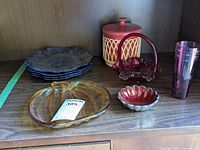 Wide view of shelf showing four blue textured salad plates stacked on left, a red and white ceramic container behind a red rose-themed basket dish, a ringed red enameled bowl, pumpkin-shaped amber glass plate, and purple tall drinking glass on right.