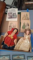 Two small dolls in a box with decorative dated fashion prints on paper behind them, showing condition and details.