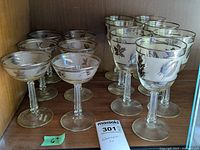 Full view of six champagne saucers and seven wine glasses with silver leaf pattern on shelf.