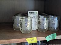 Photo of 17 silver toned leaf print juice glasses arranged in rows on a wooden shelf. The glasses show frosted bands with leaf designs and silver rims.