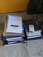 Two stacks of assorted notebooks and sketchbooks on floor with various sizes and bindings.