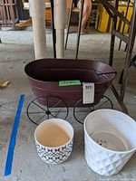 Three planters displayed together: two ceramic planters (white with black geometric pattern and plain white) and one reddish-brown metal wagon-style planter with four wire wheels.