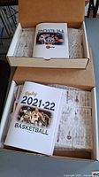 Two cardboard boxes containing Replay Publishing basketball sets from 2013-14 and 2021-22. Each set includes a rule book visible on top with basketball images on covers.