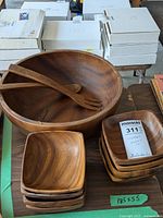 View of large wooden salad bowl with two wooden salad serving utensils inside, stacked with eight small wooden serving bowls nearby.