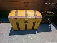 Full front and side view of yellow wooden steamer trunk with brown wooden strips and metal accents sitting on floor.