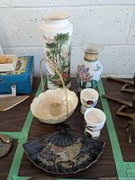 Photo of 2 ceramic Asian-themed vases, 2 egg cups with floral decorations, cream basket with candle holder and metal handle, and black fan-shaped bonbon dish with floral design.