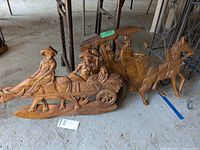 Two wooden wall hanging oxen carts with figures, placed on concrete floor with metal racks behind. Visual details show wood grain and wear.