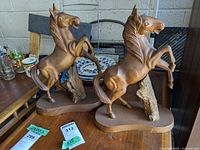 Two vintage wooden horse statues shown on a table, with some background objects including plates. Both horses are in a rearing stance on wooden bases with natural wood color and visible wear like scratches and scuffs.