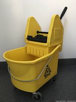 Side view of the assembled yellow mop bucket with wringer attachment, showing casters and warning label.
