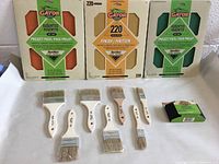 Photo of Gator sanding block, grit assorted project packs, fine finish sandpaper, and set of 7 paint brushes laid out on surface.