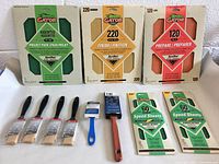 Photo showing the full lot with assorted Gator sandpaper in 80, 120, 220 grits in large sheets 9x11 inches, four small Good brand 1-inch flat paint brushes, a blue-handled 1-inch pad trimmer brush, a wooden-handled angular 2-inch Pintar brush, and two packs of Gator 60 grit speed sheets.