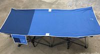 Top view of blue extra wide folding camping cot with dark blue accents, showing fabric stretched on black frame with small side pocket.