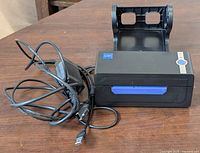 Front view of the black LH2 thermal label printer with attached power cord and plastic holder for label rolls, placed on a wooden surface.