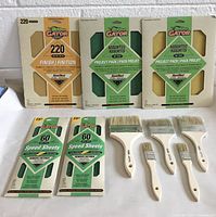 Photo showing 3 packages of assorted grit sandpaper sheets 9"x11" and 2 packages of 60 grit speed sheets 3-2/3"x9", with five paint brushes arranged in front.