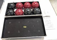 Open metal carrying case showing four red and four black bocce balls arranged inside with one white pallino ball and instruction sheet.
