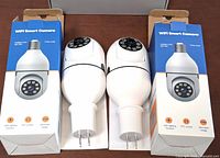 Two white WiFi smart cameras in E27 light bulb form factor displayed with their packaging boxes showing product features such as FHD image and PTZ control.