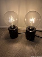 Two small Edison bulb table lamps lit and placed on a wooden floor against a plain light wall, showing filament bulbs and black cylindrical bases.