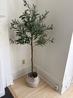 Full view of the artificial olive tree plant standing in the corner of a room on wood floors. The tree is in a white and brown woven basket with handles.