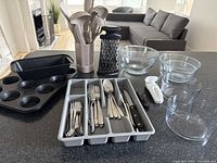 Overview of all baking items and cutlery set on kitchen counter: loaf pan, muffin tin, glass bowls, basket with utensils, box grater, cutlery tray with flatware and kitchen knife