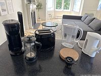 Black drip coffee maker, French press, 2-slice toaster, water filter pitcher, cork coasters, coffee capsule holder, and white electric kettle on kitchen counter and table.
