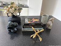 Display on dark countertop of decorative items including a black matte vase with artificial flowers, stone decor pieces, brass arrow sculpture, and stack of four books.